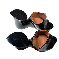 Black patent leather Alaia high-heeled mule shoes with brown insoles on a white background, featuring the brand 'Alaia' top view.