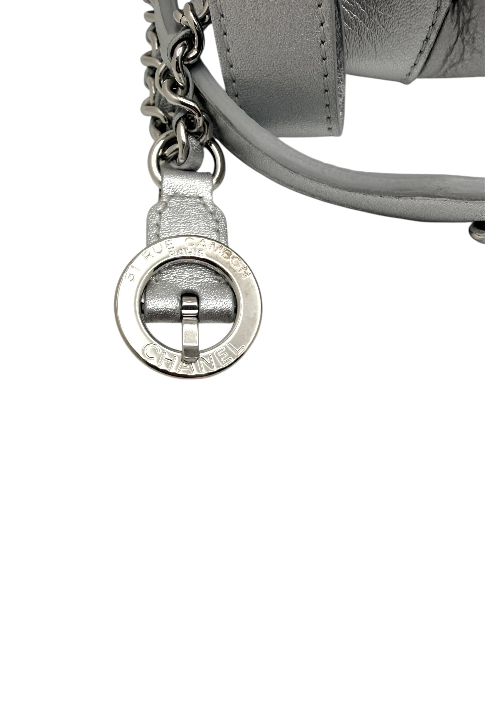 Close up of Chanel metallic silver bum bag showing engraved Rue Cambon Paris circular hardware charm with leather and chain strap attachment.