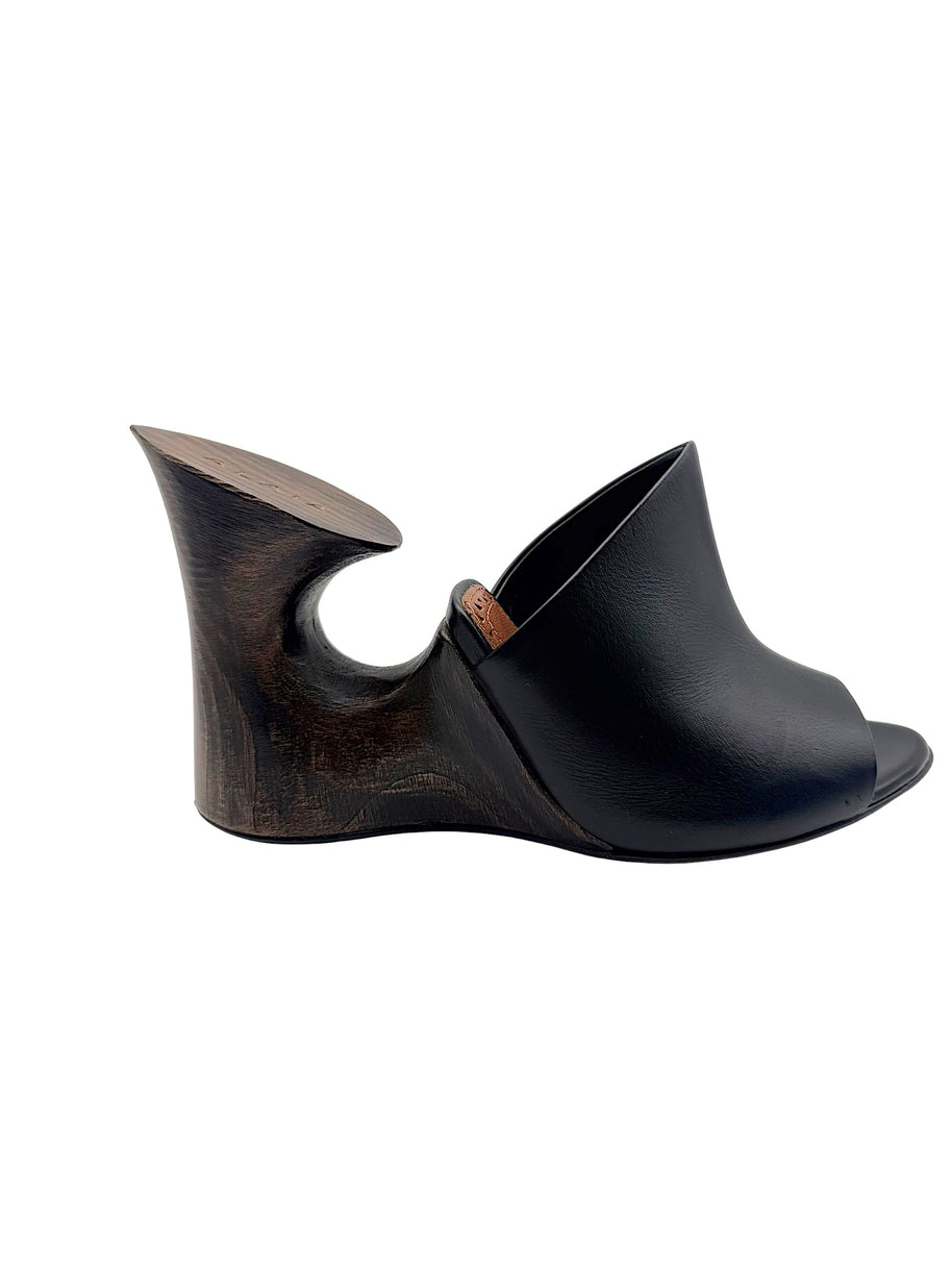 Side profile of an Alaïa La Sculpture black leather mule showing its architectural wooden wedge heel with circular cutout.
