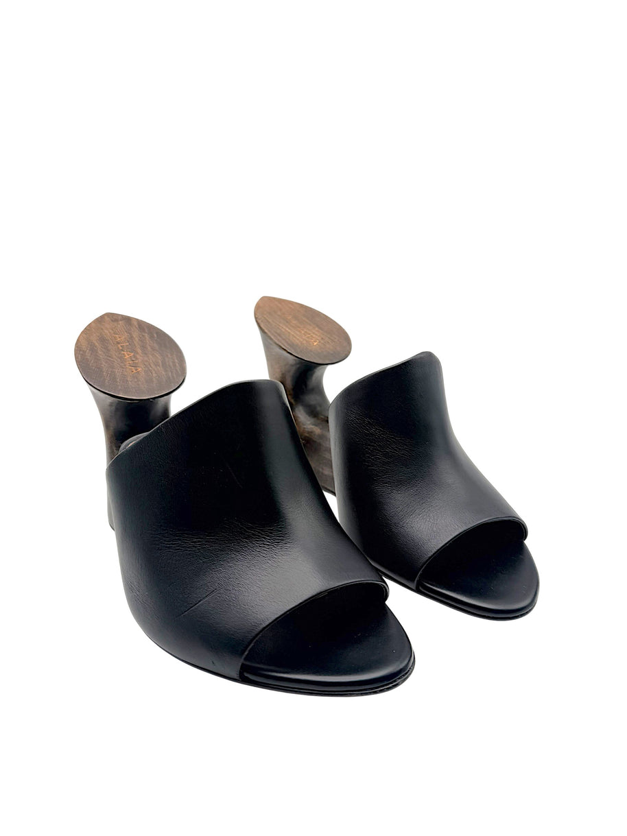 Angled front view of Alaïa La Sculpture black leather mules with wide leather uppers, open toes and sculptural wooden wedge heels.