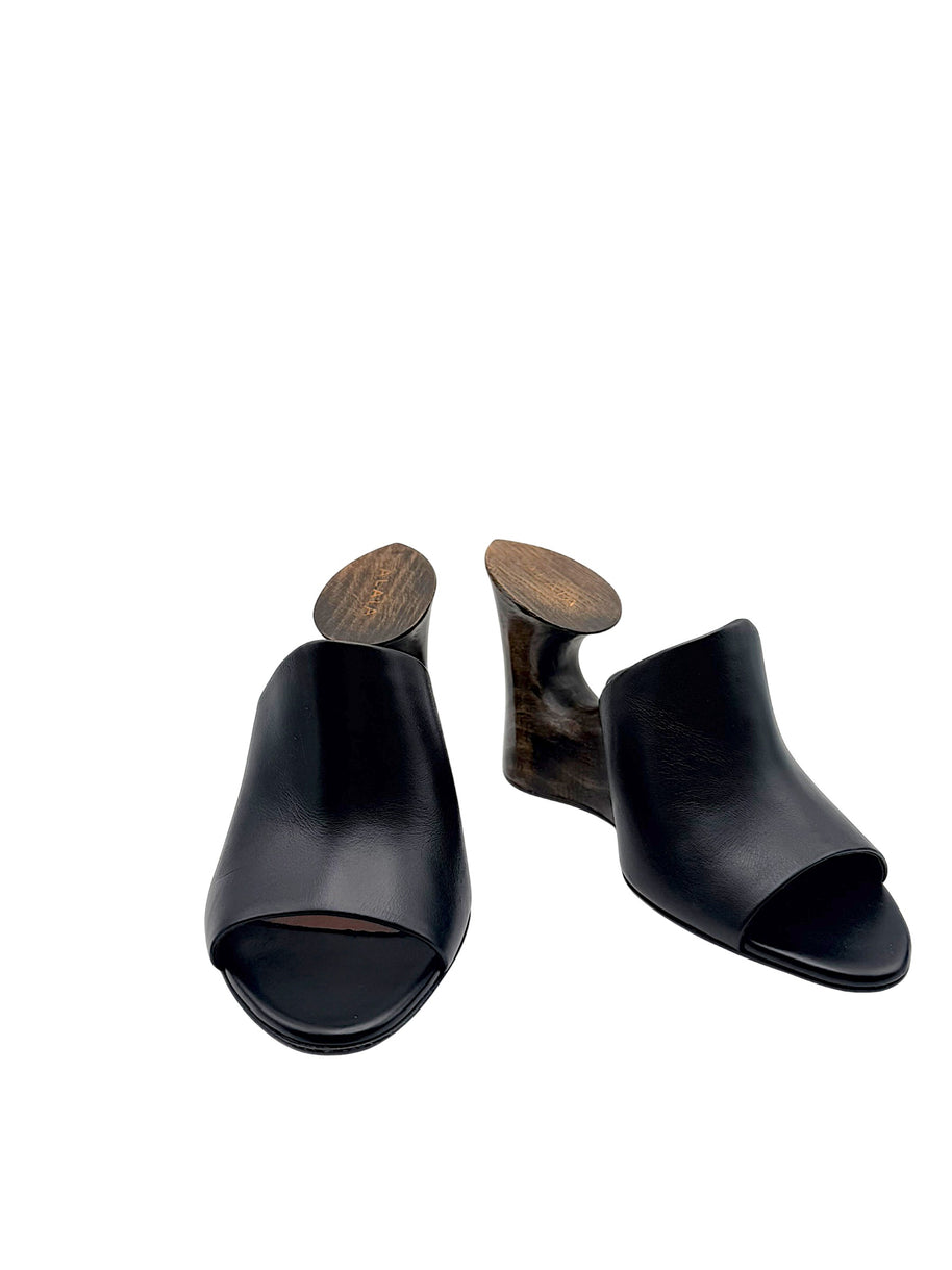 Alaïa La Sculpture black leather open-toe mules in front three-quarter view, featuring architectural dark wood cutout wedge heels.