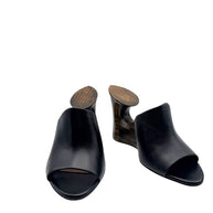 Alaïa La Sculpture black leather open-toe mules in front three-quarter view, featuring architectural dark wood cutout wedge heels.