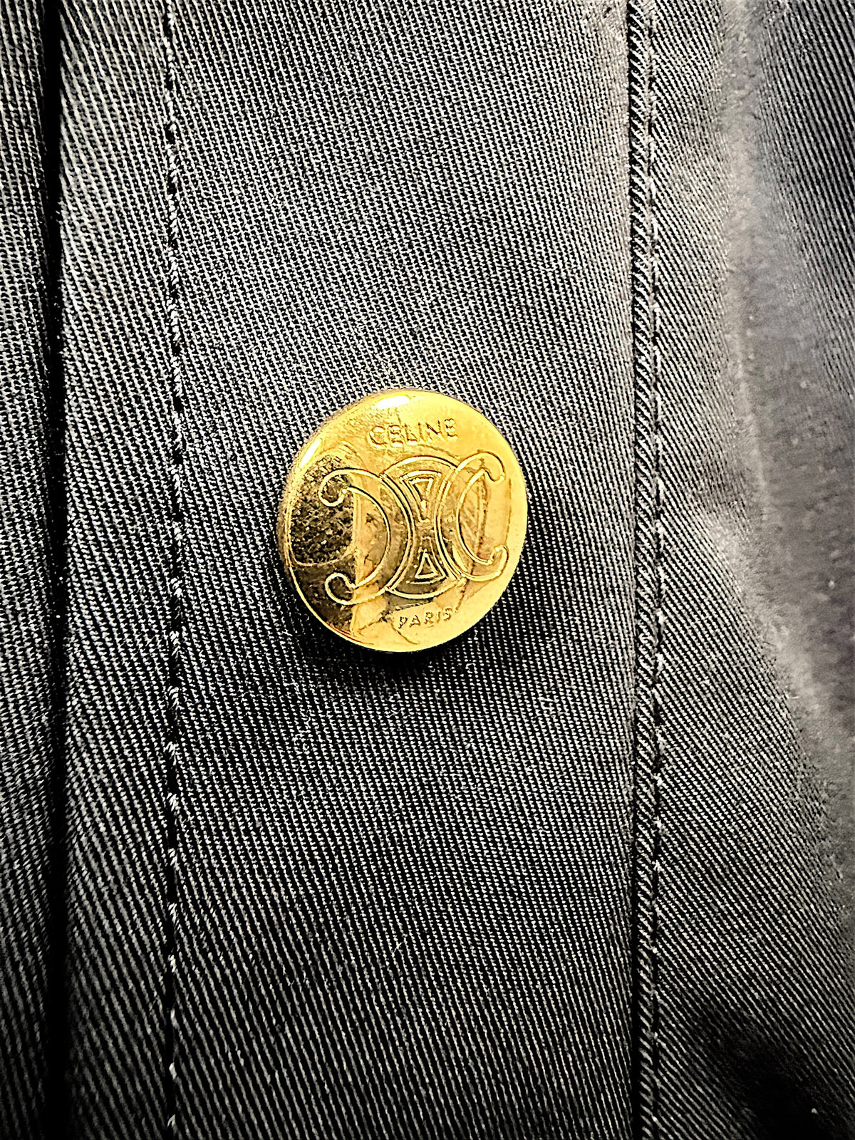 Close-up of gold-tone Celine Paris Triomphe logo button on black cotton skirt, highlighting branded hardware and fabric texture.