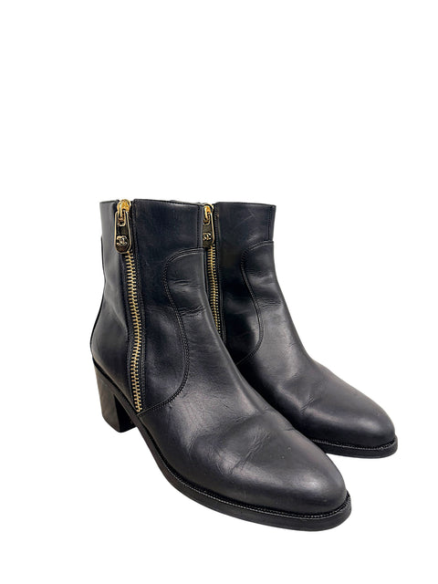 Front three-quarter view of Chanel black leather ankle boots with gold-tone CC zipper pulls, curved leather panels and stacked block heels.