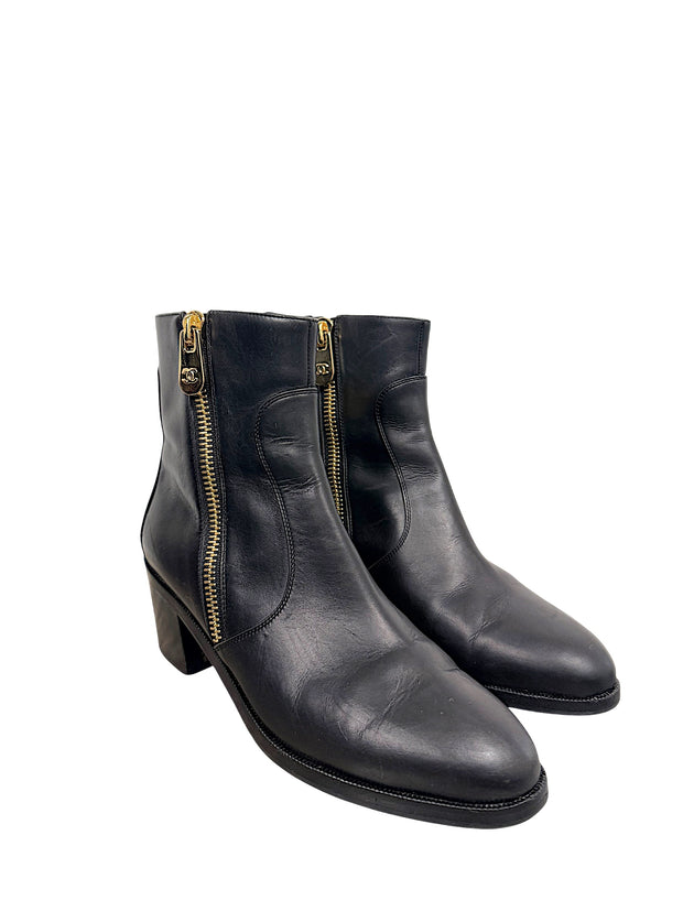 Front three-quarter view of Chanel black leather ankle boots with gold-tone CC zipper pulls, curved leather panels and stacked block heels.