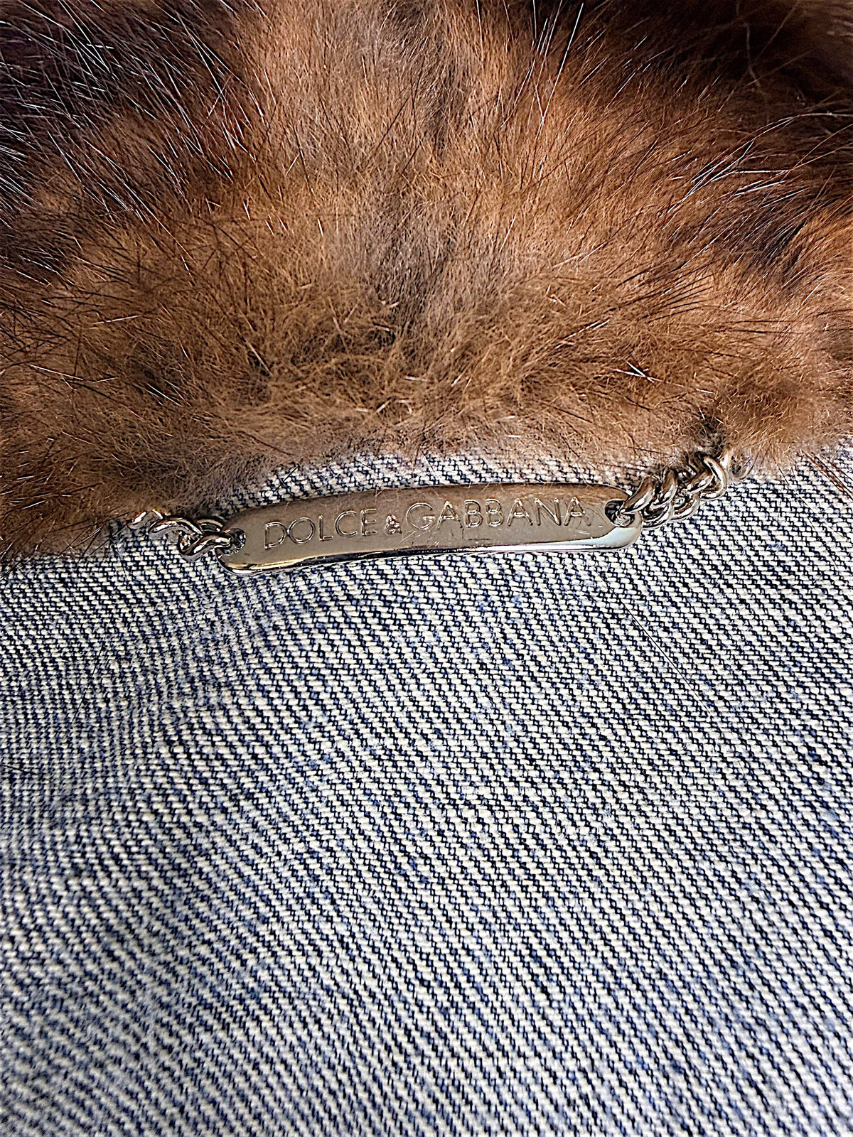 Close-up of Dolce & Gabbana engraved silver-tone metal chain logo plaque inside vintage blue denim jacket beneath the plush brown fur collar.