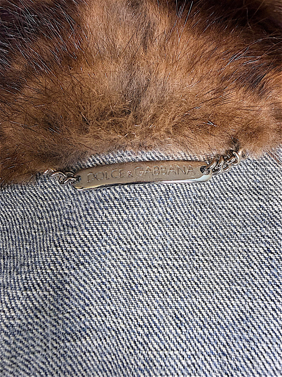 Close-up of Dolce & Gabbana engraved silver-tone metal chain logo plaque inside vintage blue denim jacket beneath the plush brown fur collar.