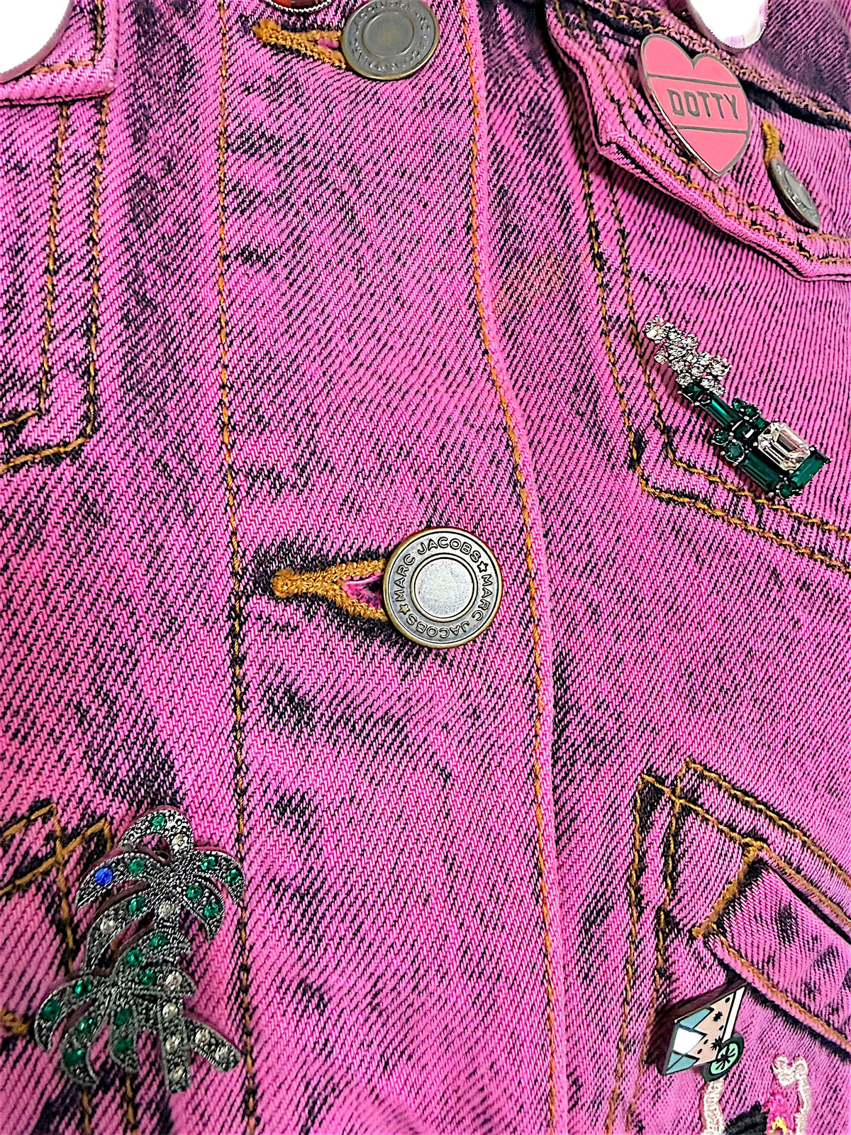 Detailed view of Marc Jacobs engraved metal buttons, pink DOTTY heart pin, palm tree embellishment and acid wash denim stitching.