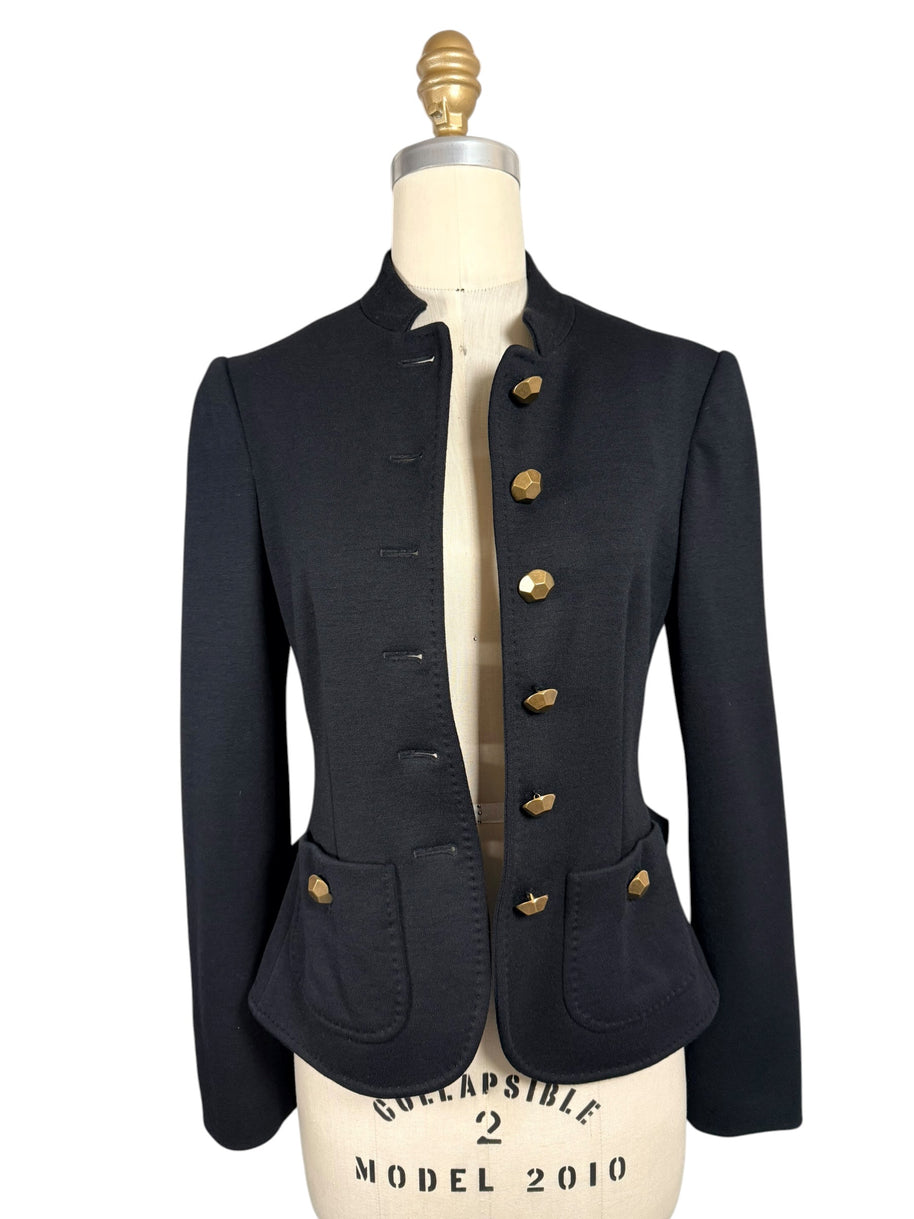 Moschino black tailored peplum jacket shown unbuttoned on a dress form, featuring a stand collar, faceted gold-tone buttons and rounded front pockets.
