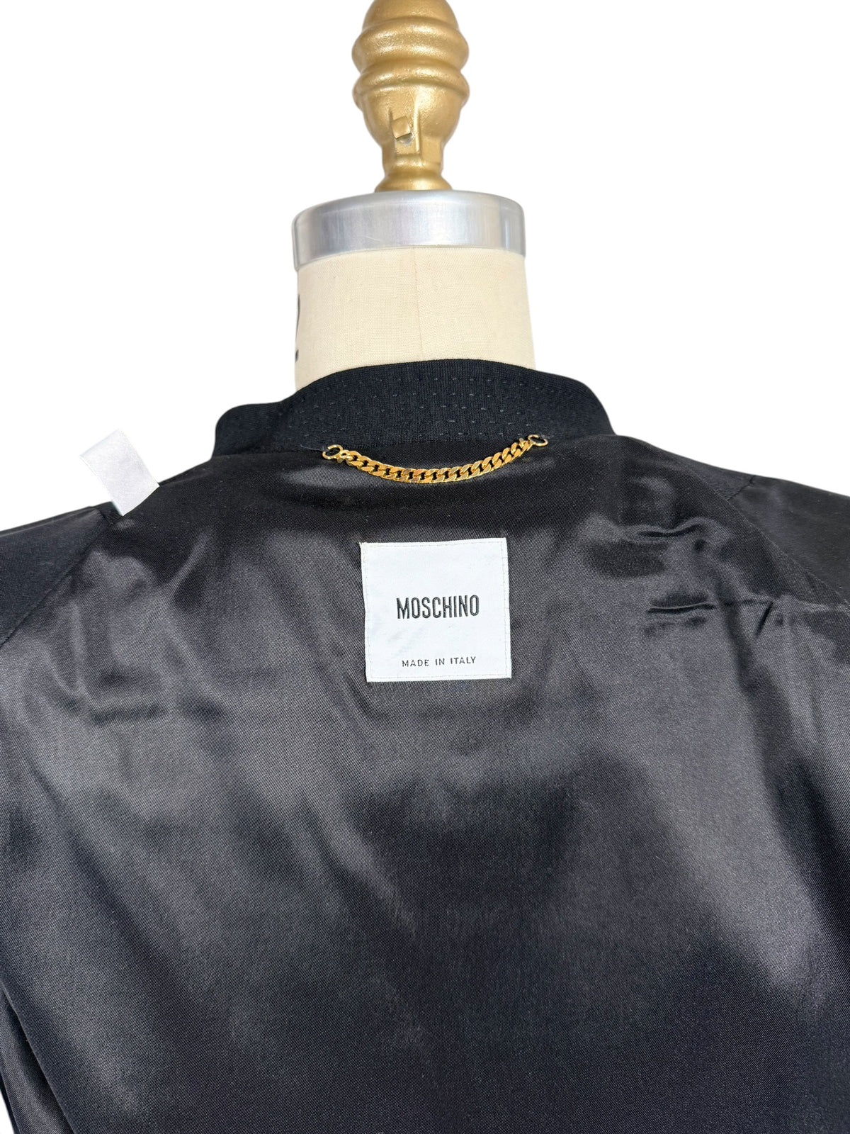 Close-up of Moschino Made in Italy designer label inside black peplum jacket, showing the black satin lining, textured collar and gold-tone hanging chain.