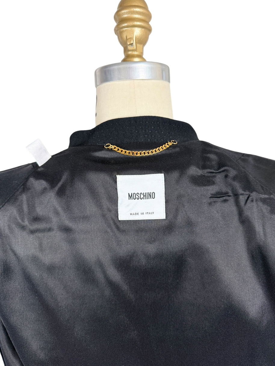Close-up of Moschino Made in Italy designer label inside black peplum jacket, showing the black satin lining, textured collar and gold-tone hanging chain.