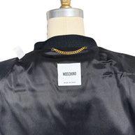 Close-up of Moschino Made in Italy designer label inside black peplum jacket, showing the black satin lining, textured collar and gold-tone hanging chain.