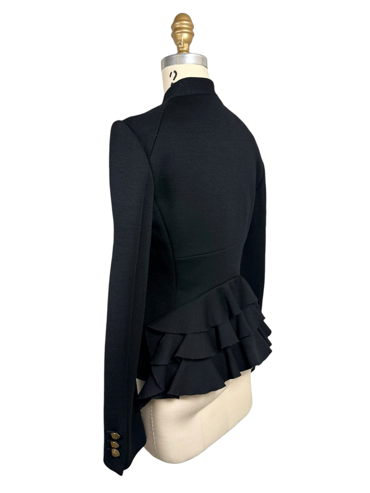 Three-quarter rear view of Moschino black tailored jacket highlighting the sculptural layered ruffle peplum, contoured back seams and gold-button cuff.