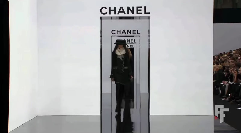 14-second runway video of Chanel Fall 2009 Runway Look 22 under Karl Lagerfeld, featuring the original black bouclé tweed jacket and matching skirt with ivory fringe trim, emerald green accents, oversized tweed hat, and ivory fur collar as presented on the Paris runway.