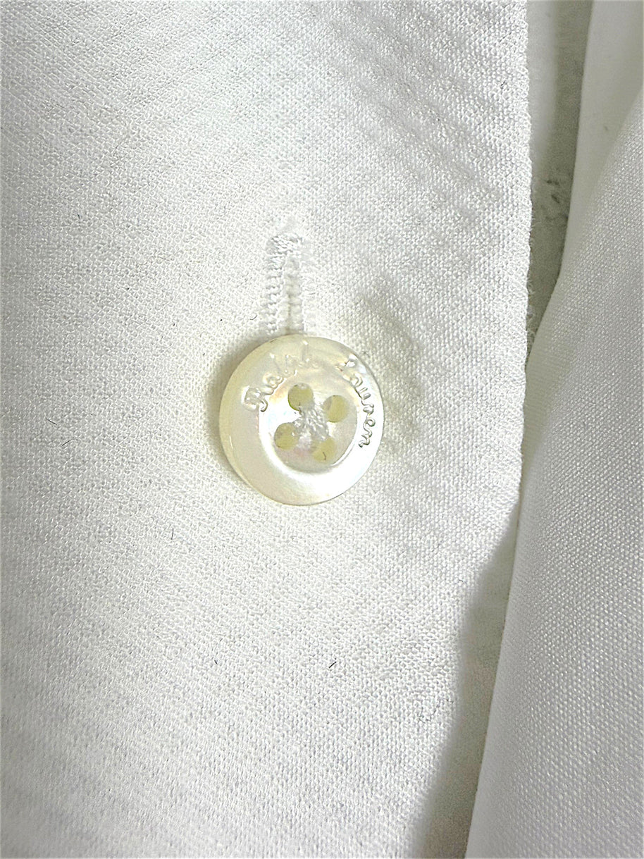 Macro detail of a lustrous Ralph Lauren engraved button on the white cotton tuxedo shirt, showing fabric texture and stitching.