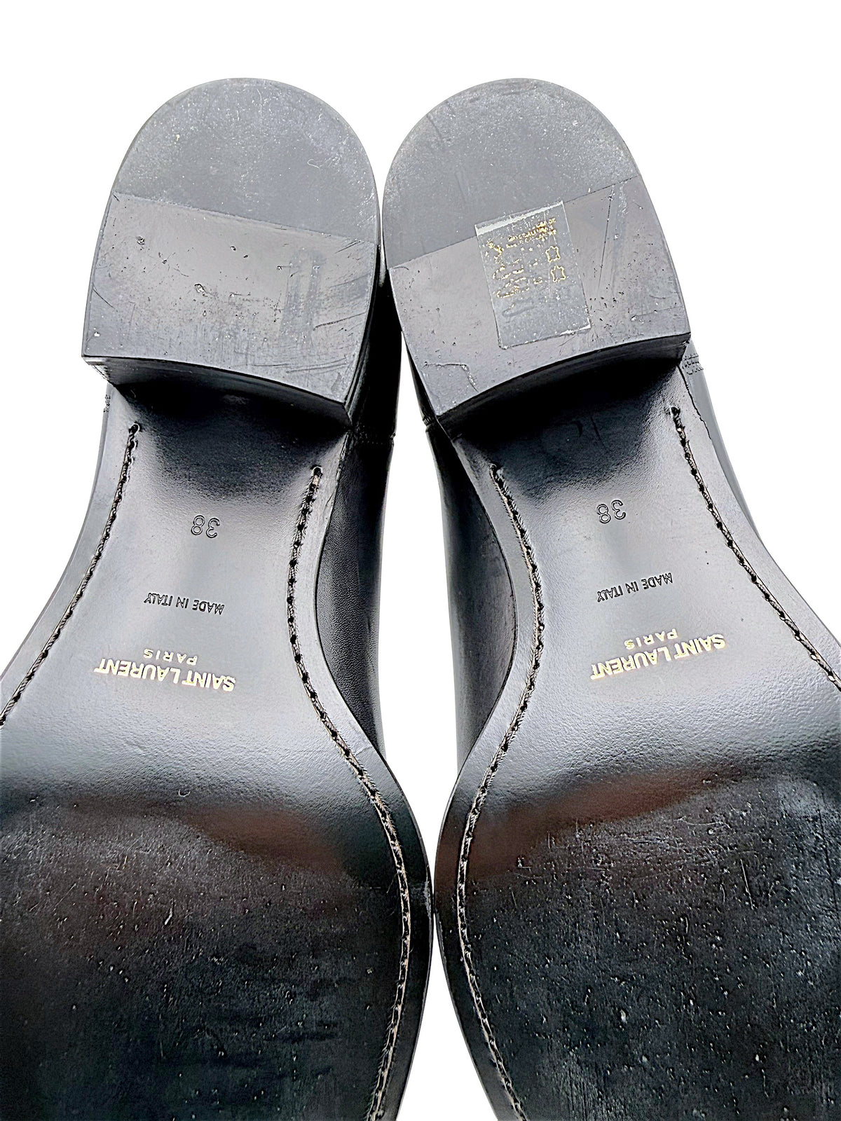 Detailed view of Saint Laurent Paris black leather Chelsea boot soles showing gold designer logos, Made in Italy stamps, European size 38 and surface wear on stacked heels and leather outsoles.