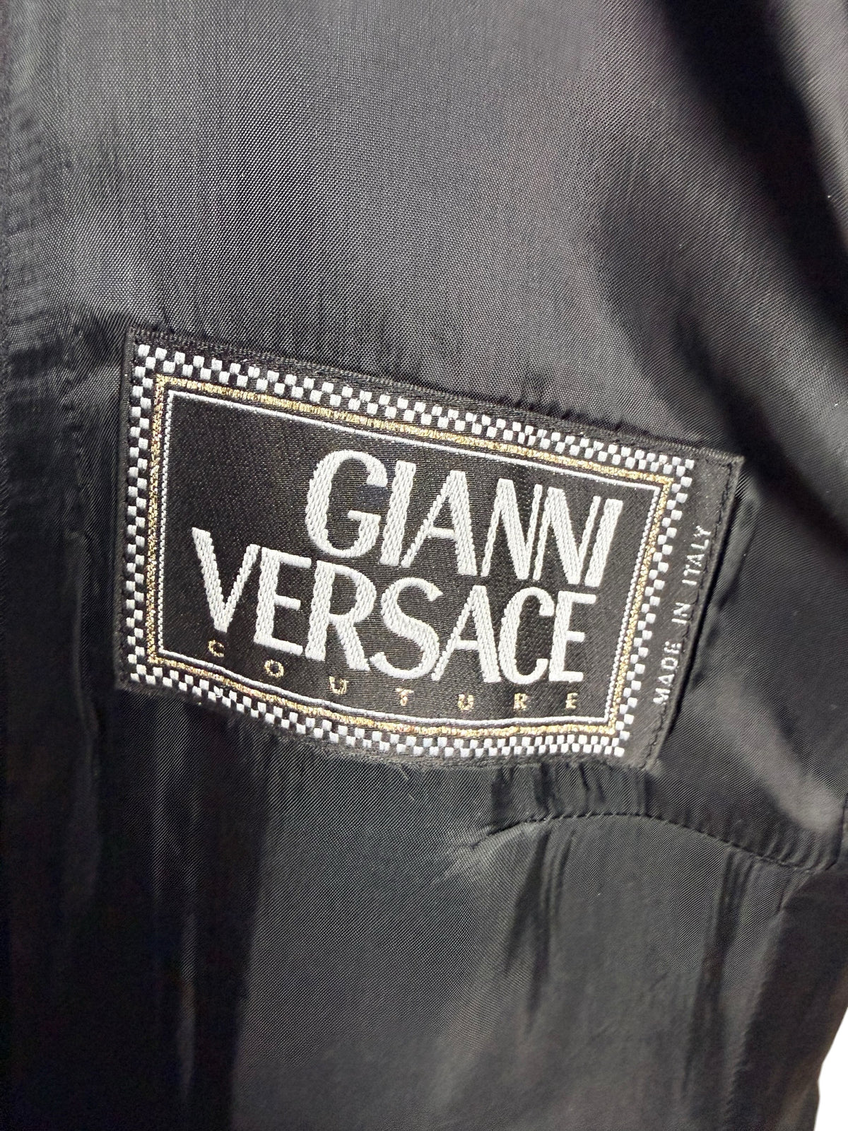 Close-up of the original Gianni Versace Couture woven designer label with black and white checkered border, gold detailing and Made in Italy marking inside a vintage black blazer.